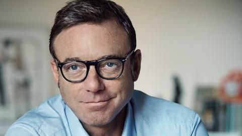 Schmunzelnd blickt ein Mann mit runder Brille und hellblauem Hemd in die Kamera. Er hat kurzes, braunes Haar, im Hintergrund sieht man verschwommen ein Arbeitszimmer.