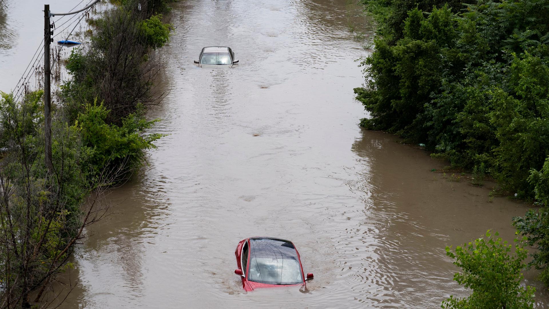 Zu sehen sind mehrere Autos, die auf einer überfluteten Straße zu einem Großteil unter Wasser stehen. 