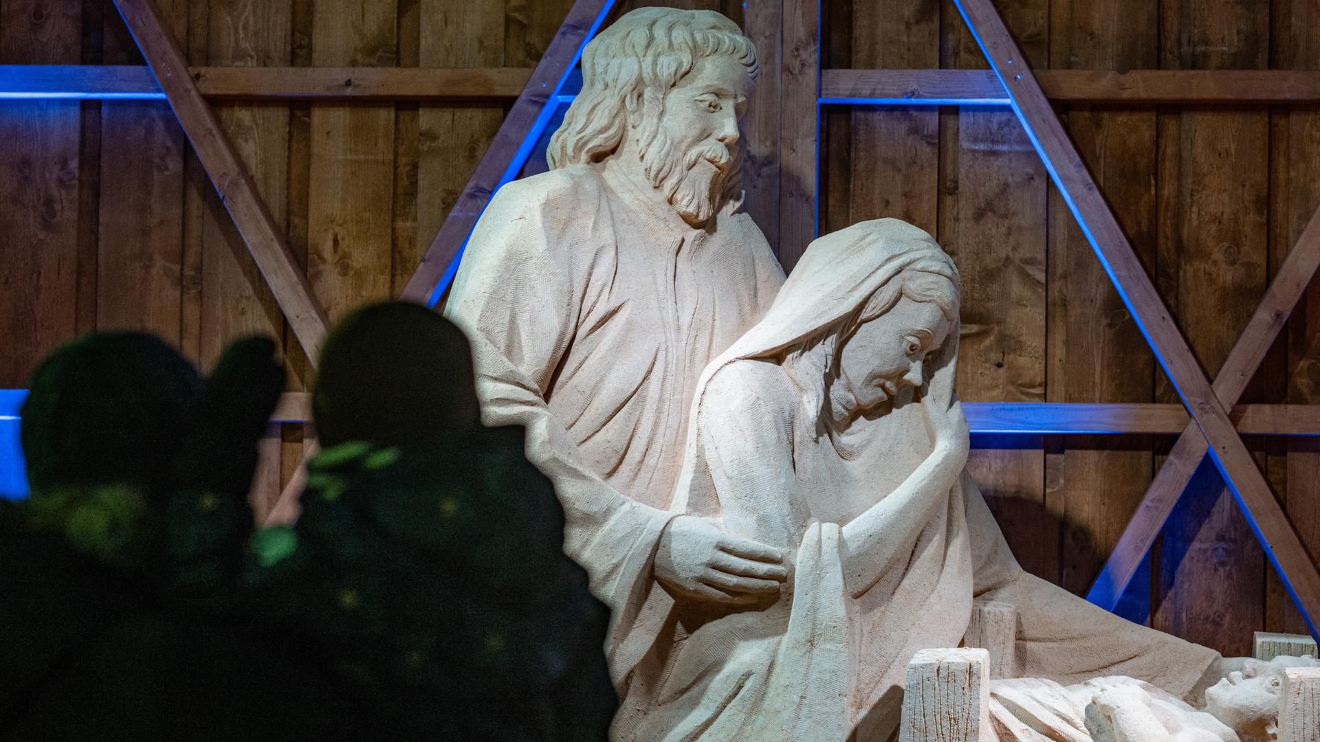 Der Bildausschnitt zeigt Maria, Josef und das Jesuskind. Die hölzerne Rückwand ist zum Teil blau angestrahlt. Im Vordergrund links sind verschwommen Besucher zu sehen. Der Bildausschnitt zeigt Maria, Josef und das Jesuskind. Die hölzerne Rückwand ist zum Teil blau angestrahlt. Im Vordergrund links sind verschwommen Besucher zu sehen.