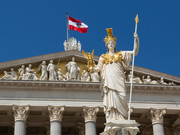 Die Statue der Pallas Athene steht vor dem Parlament in Wien, hinter ihr flattert die österreichische Flagge im Wind.