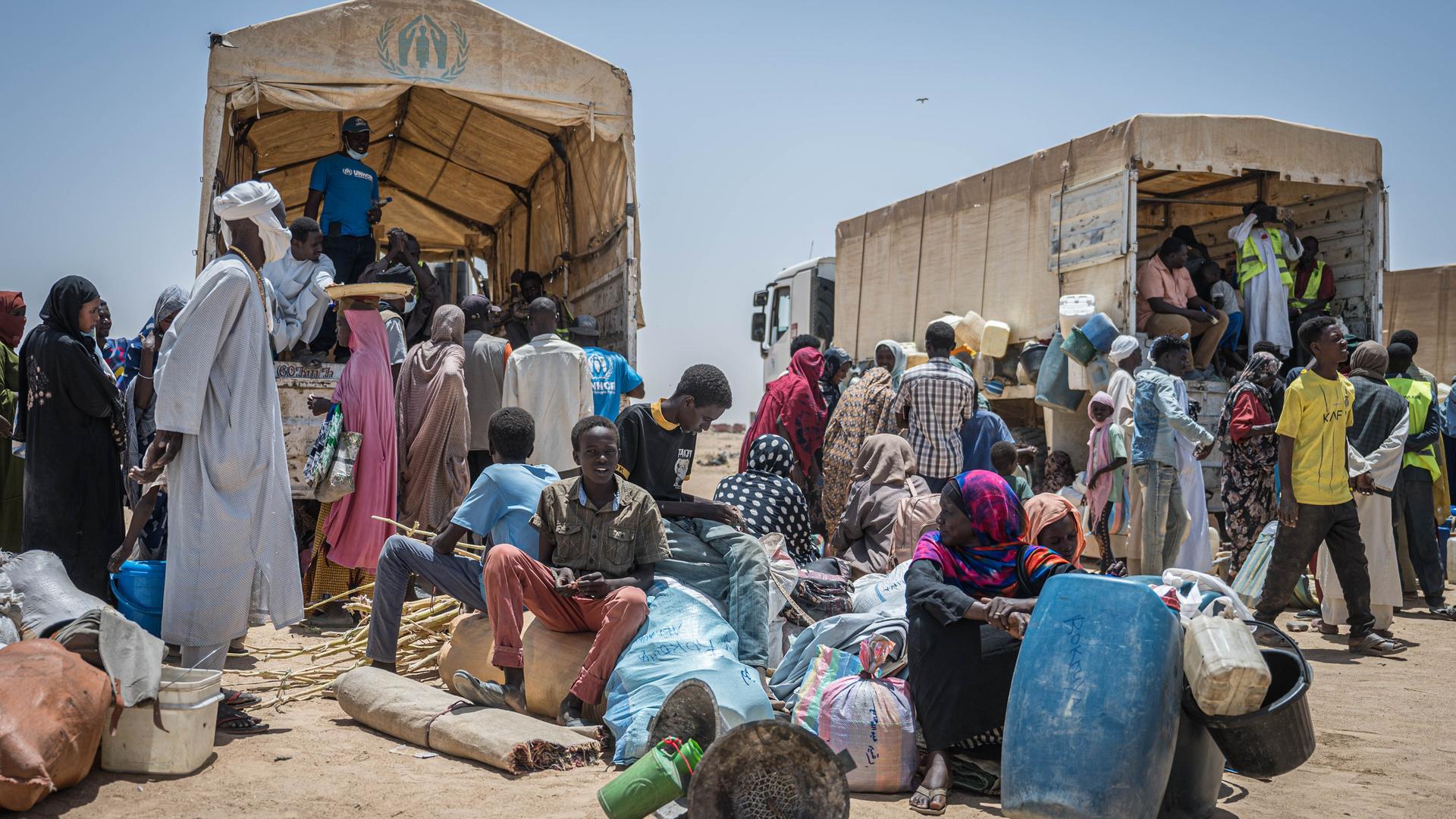 Menschen im Sudan in traditioneller Kleidung sitzen vor zwei LKW und scheinen zu warten