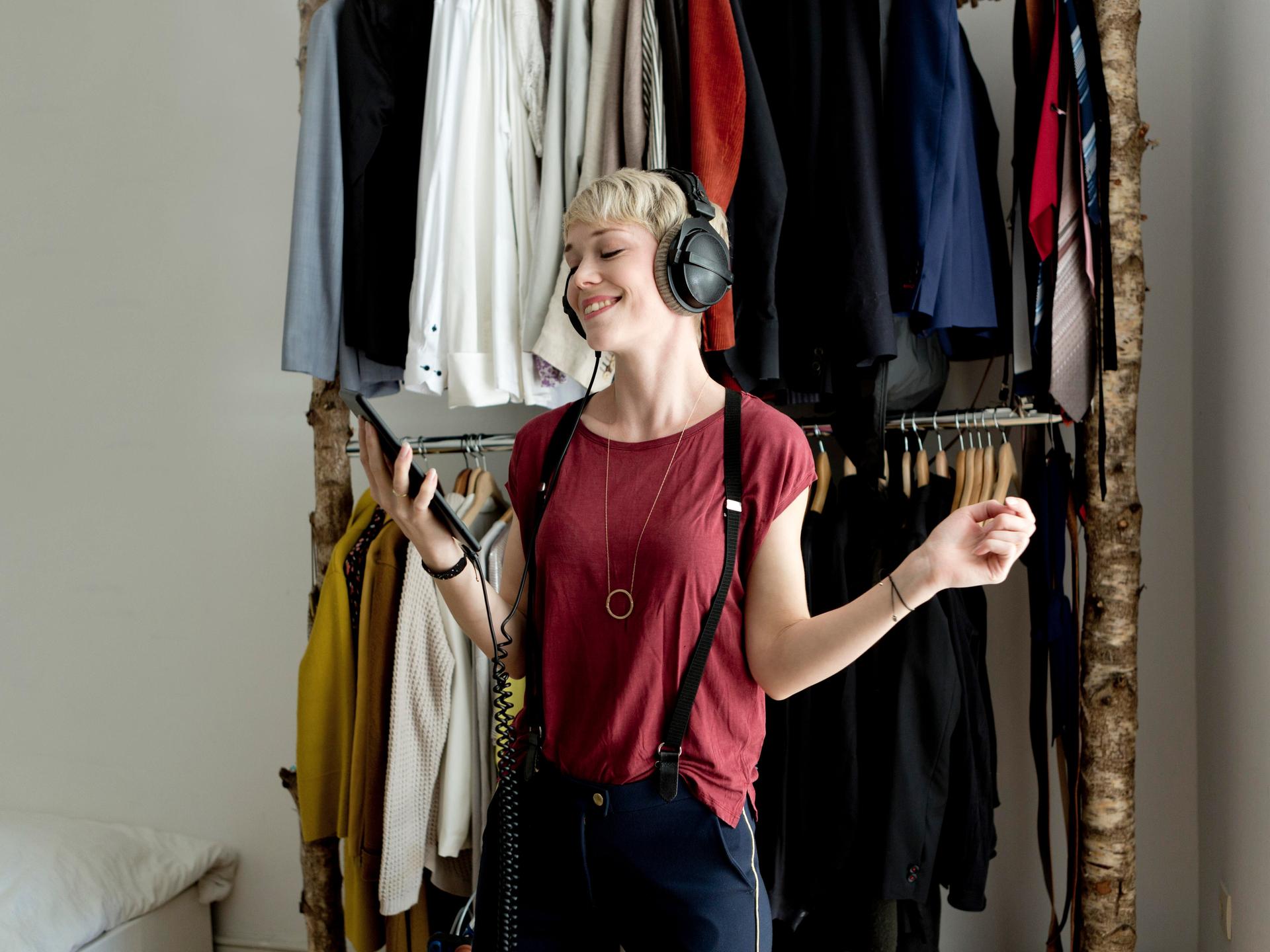 Eine junge Frau hört mit Kopfhörern Musik von ihrem Smartphone. Im Hintergrund ist eine Kleiderstange mit vielen Kleidungsstücken zu sehen.