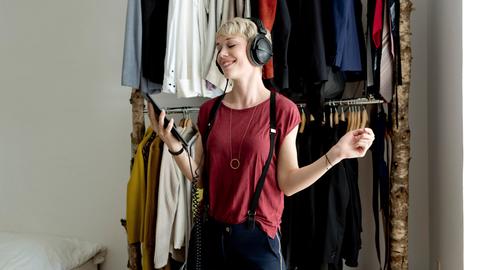 Eine junge Frau hört mit Kopfhörern Musik von ihrem Smartphone. Im Hintergrund ist eine Kleiderstange mit vielen Kleidungsstücken zu sehen.