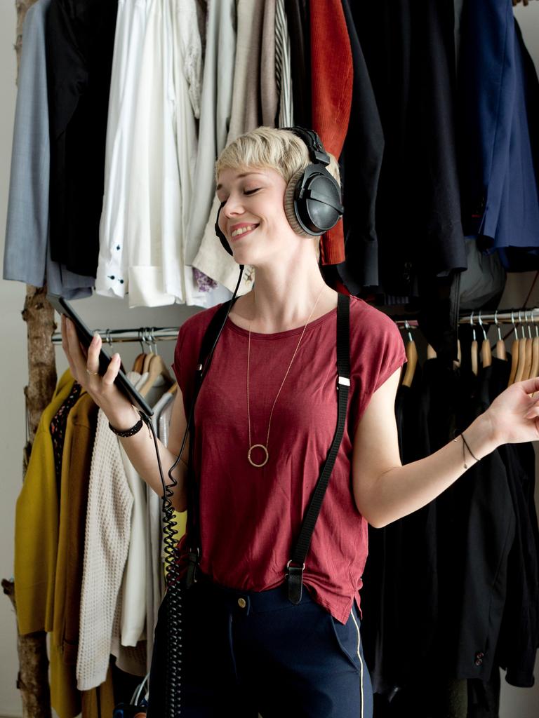 Eine junge Frau hört mit Kopfhörern Musik von ihrem Smartphone. Im Hintergrund ist eine Kleiderstange mit vielen Kleidungsstücken zu sehen. Eine junge Frau hört mit Kopfhörern Musik von ihrem Smartphone. Im Hintergrund ist eine Kleiderstange mit vielen Kleidungsstücken zu sehen.