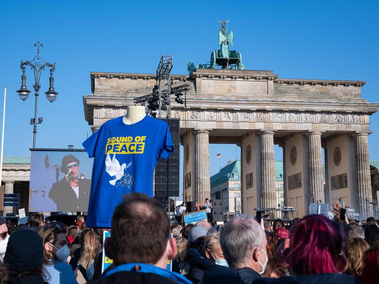 Zuschauer vor dem Brandenburger Tor verfolgen das Geschehen bei der Friedensaktion "Sound of Peace", während Marius Müller-Westernhagen auf einem Bildschirm zu sehen ist.