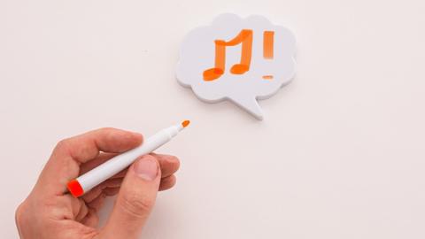 Eine linke Hand hat auf einen Klebezettel zwei orangefarbene Noten gemalt.