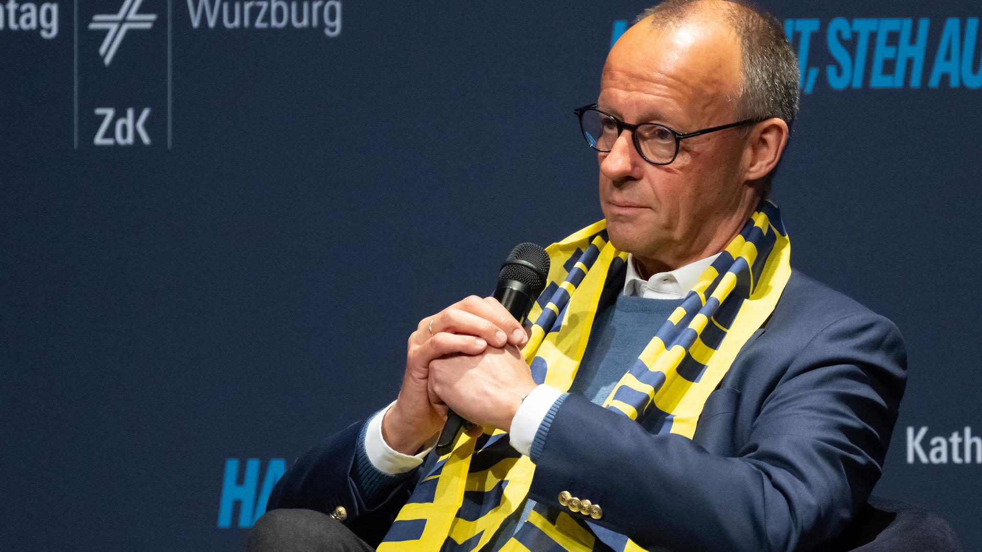 Bundeskanzler Merz sitzt mit einem Mikrofon in der Hand auf der Bühne des Deutschen Katholikentags in Würzburg.