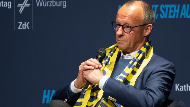 Bundeskanzler Merz sitzt und hat ein Mikrofon in der Hand. Um den Hals trägt er den gelben Schal des Katholikentags.