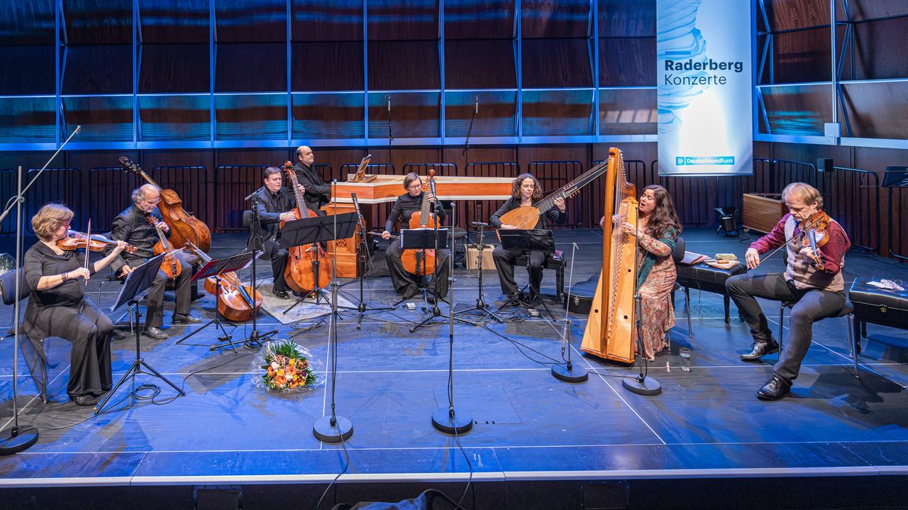 Arianna Savall und Petter Udland Johansen sitzen mit dem Ensemble Armonico Tributo Consort im Halbkreis auf einer Bühne, auf der auch ein Aufsteller mit der Schrift "Raderbergkonzert" steht.