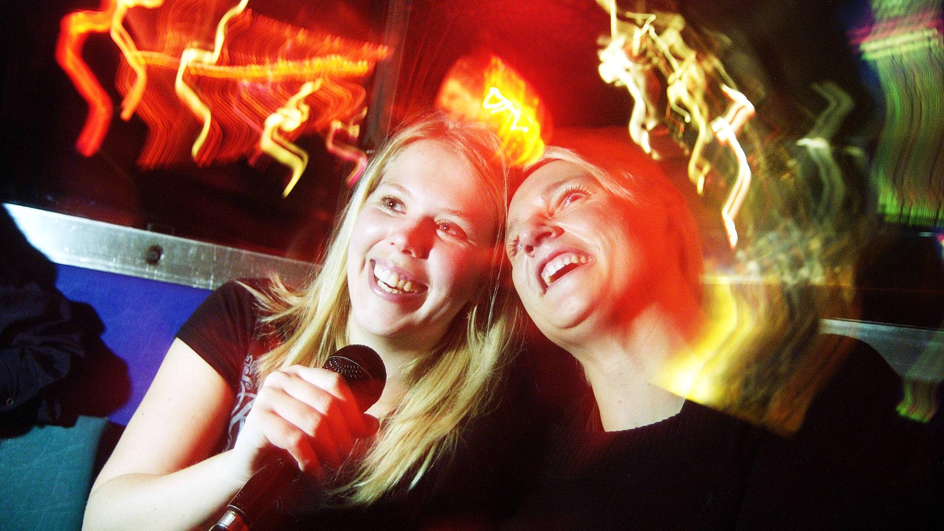 Zwei junge Frauen singen gemeinsam Karaoke in einer Bar bei entsprechend gedämpftem Licht. Sie lachen und haben offensichtlich Spaß. 