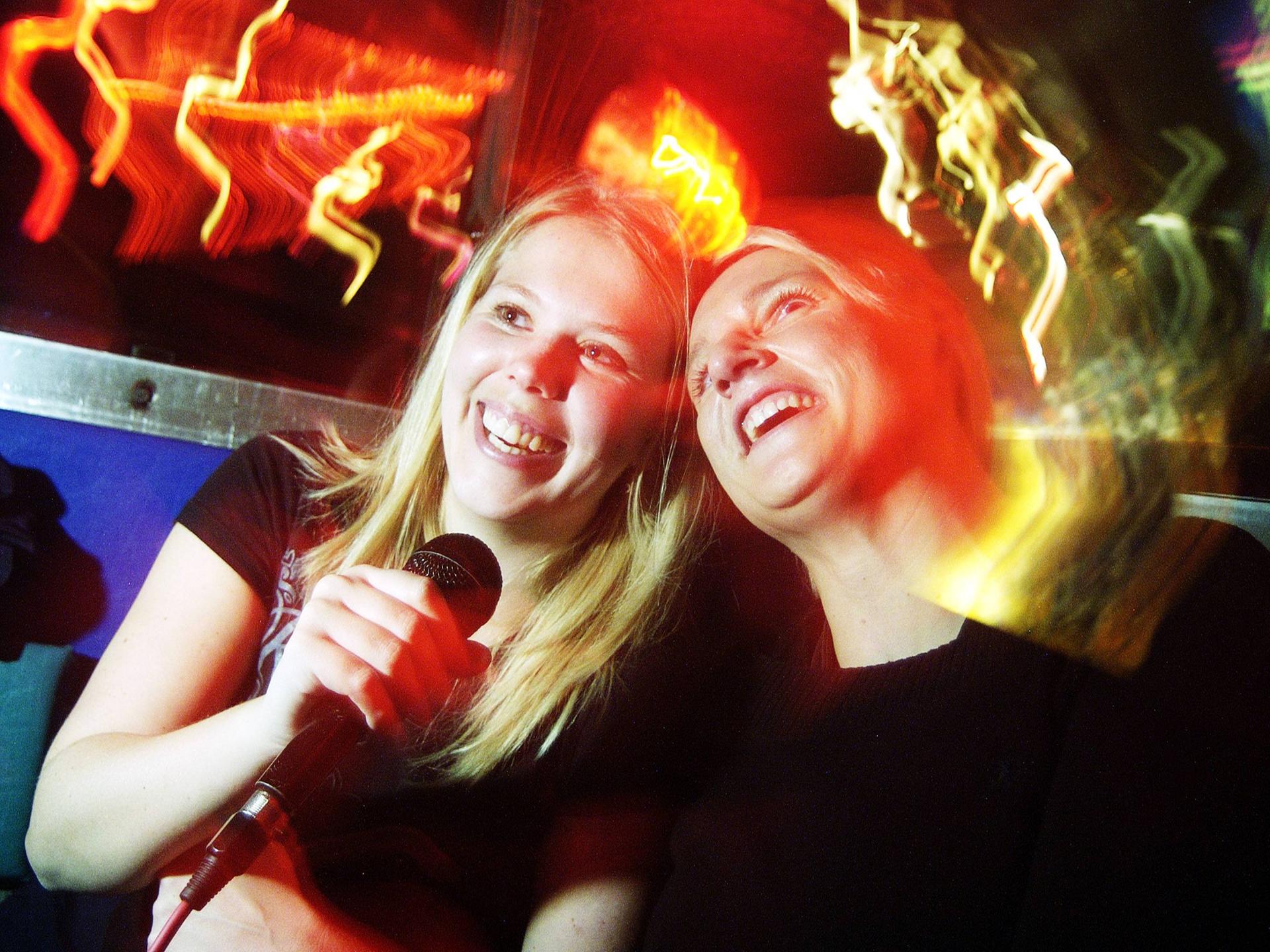 Zwei junge Frauen singen gemeinsam Karaoke in einer Bar bei entsprechend gedämpftem Licht. Sie lachen und haben offensichtlich Spaß. 