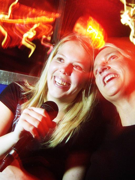 Zwei junge Frauen singen gemeinsam Karaoke in einer Bar bei entsprechend gedämpftem Licht. Sie lachen und haben offensichtlich Spaß. 