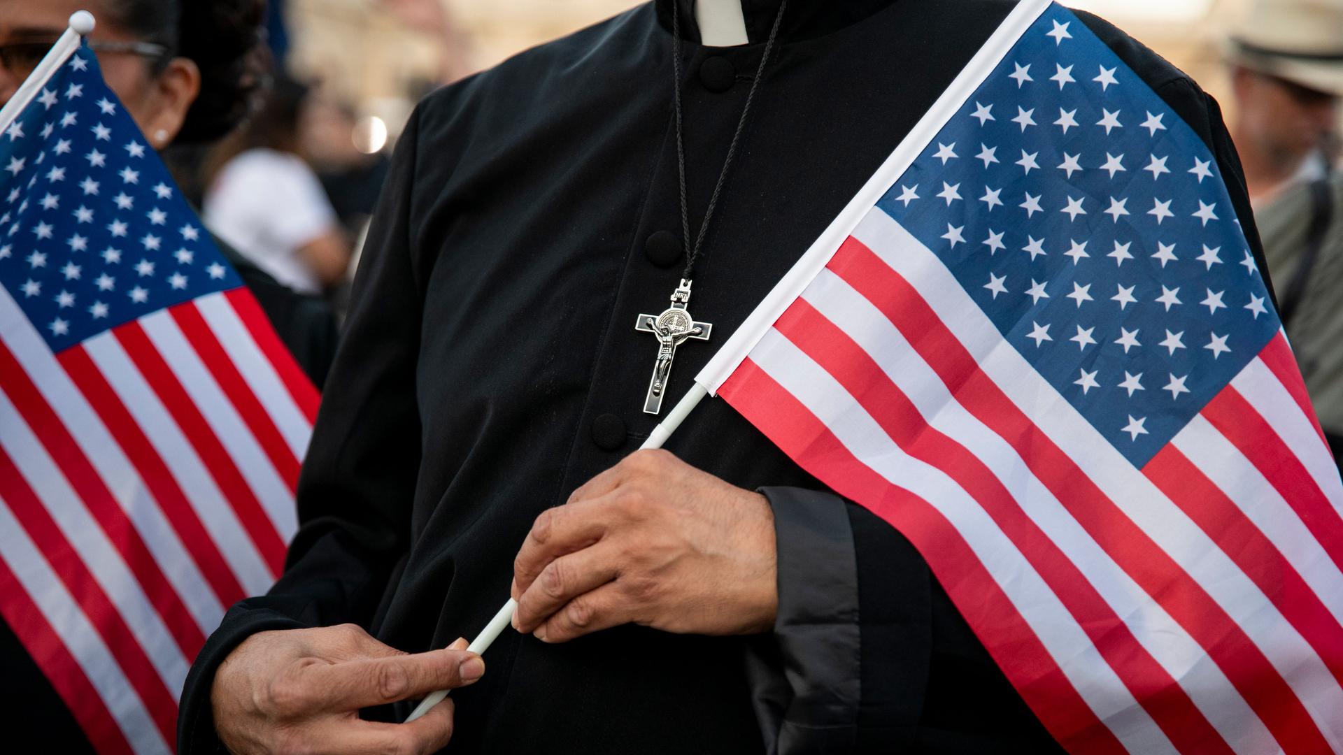 Ein Pfarrer hält eine US-amerikanische Flagge in der Hand. Auf seiner Brust ist ein silbernes Kreuz.