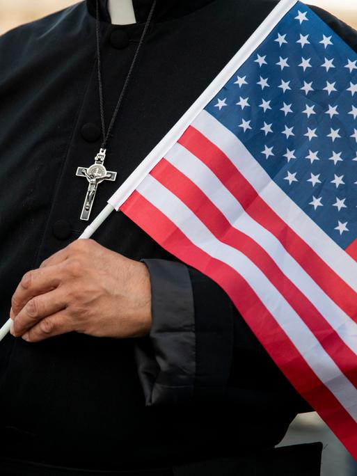 Ein Pfarrer hält eine US-amerikanische Flagge in der Hand. Auf seiner Brust ist ein silbernes Kreuz.