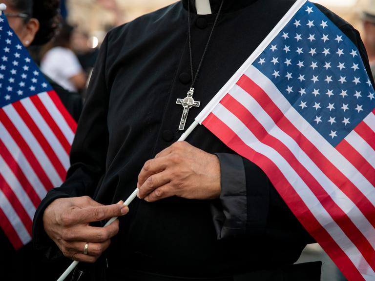 Ein Pfarrer hält eine US-amerikanische Flagge in der Hand. Auf seiner Brust ist ein silbernes Kreuz.