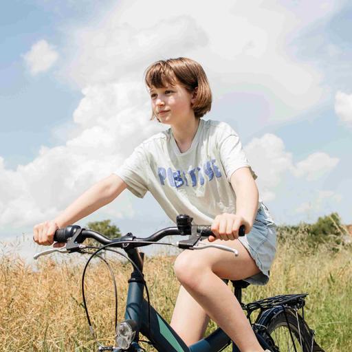 Ein Mädchen fährt unter blauem Himmel mit Wolken auf einem Fahrrad über einen Feldweg neben Getreidefeldern.