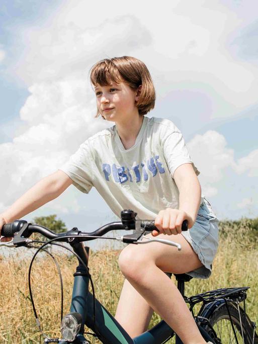 Ein Mädchen fährt unter blauem Himmel mit Wolken auf einem Fahrrad über einen Feldweg neben Getreidefeldern.