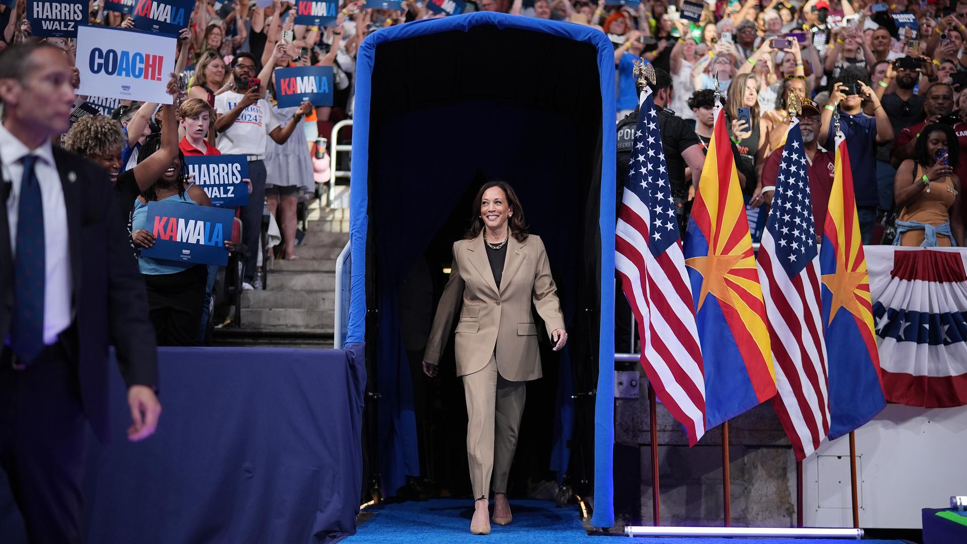 Die demokratische Präsidentschaftskandidatin und US-Vizepräsidentin Kamala Harris geht durch ein blaues Tor bei einer Wahlkampfveranstaltung in Glendale, Arizona. Um sie herum Fahnen und Fans, 9. August 2024.