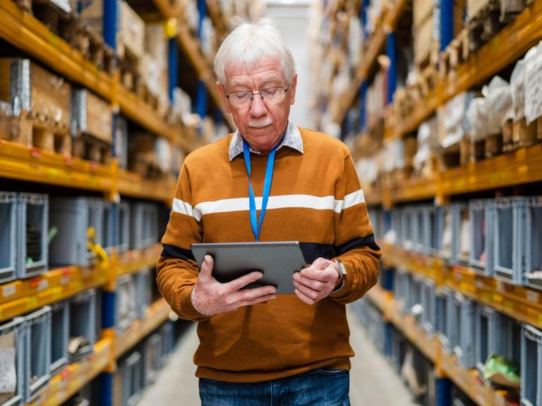 Ein älterer Mann nutzt ein Tablet während er einen Gang aus hohen Warenregalen durchgeht.