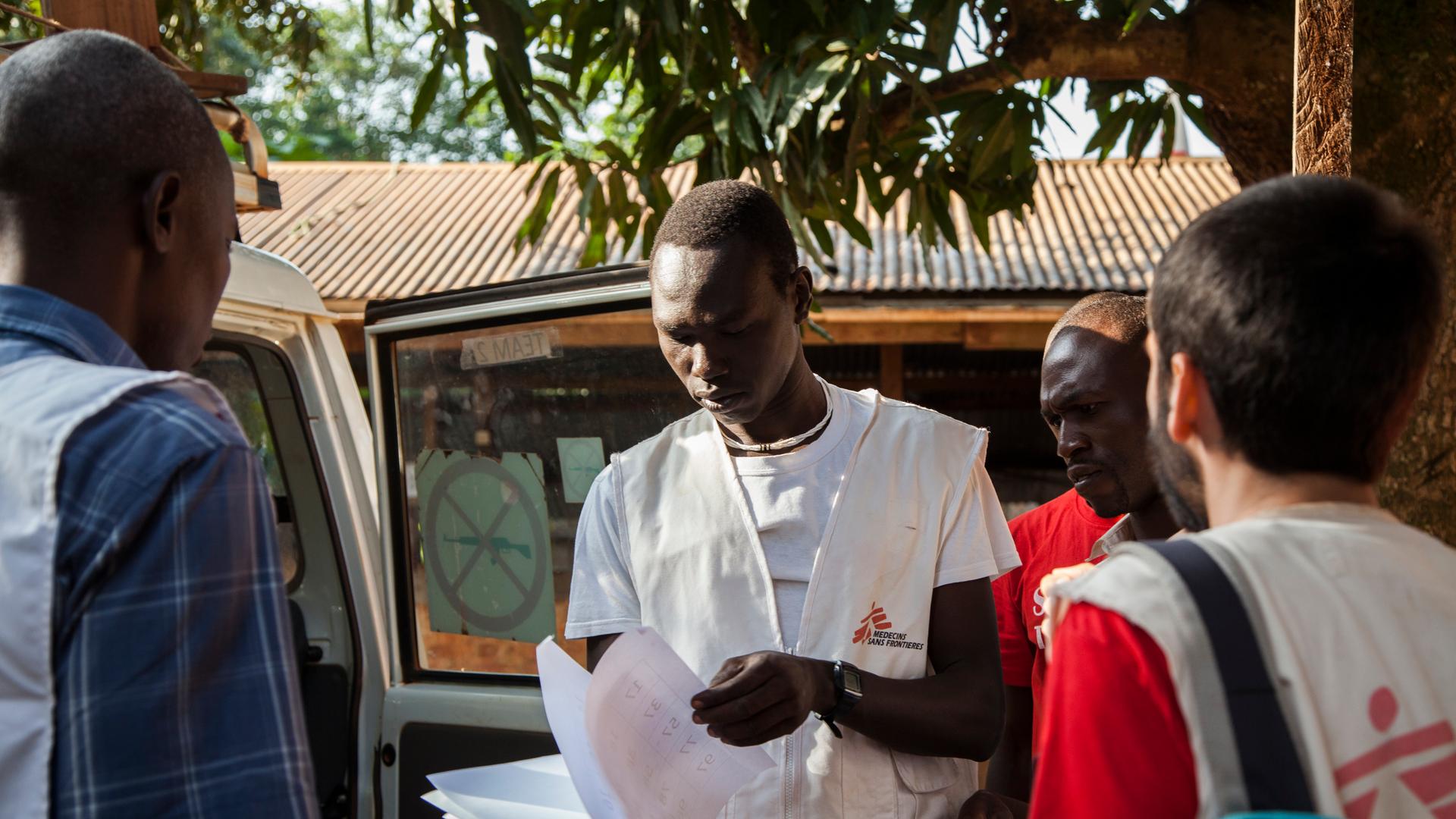 Ein junger afrikanischer Mann mit weißer Weste und "Medicins sans frontières"-Logo steht von mehreren Personen umringt neben einem Transporter und blätter in Papieren, die er in der Hand hält.