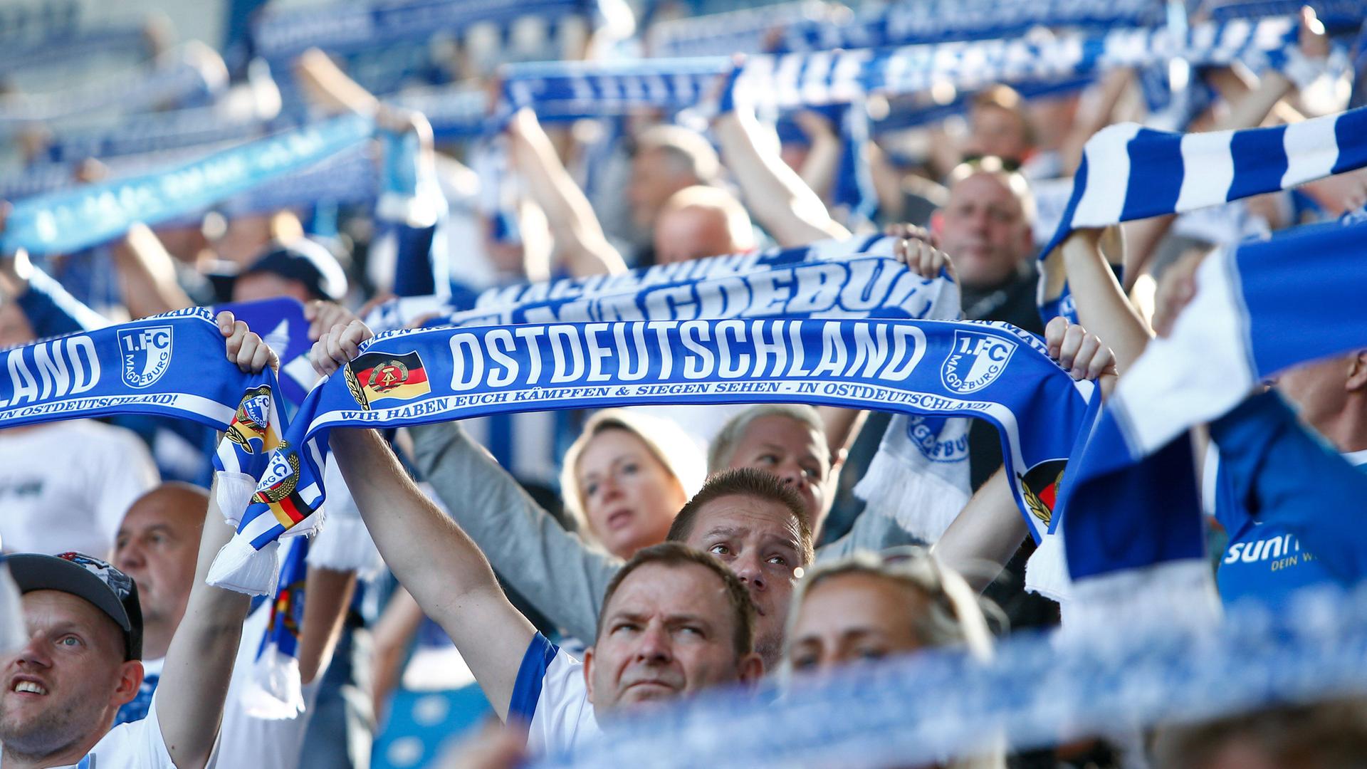 Fans des Vereins FC Magdeburg halten Schals in die Höhe mit der Aufschrift "Ostdeutschland".