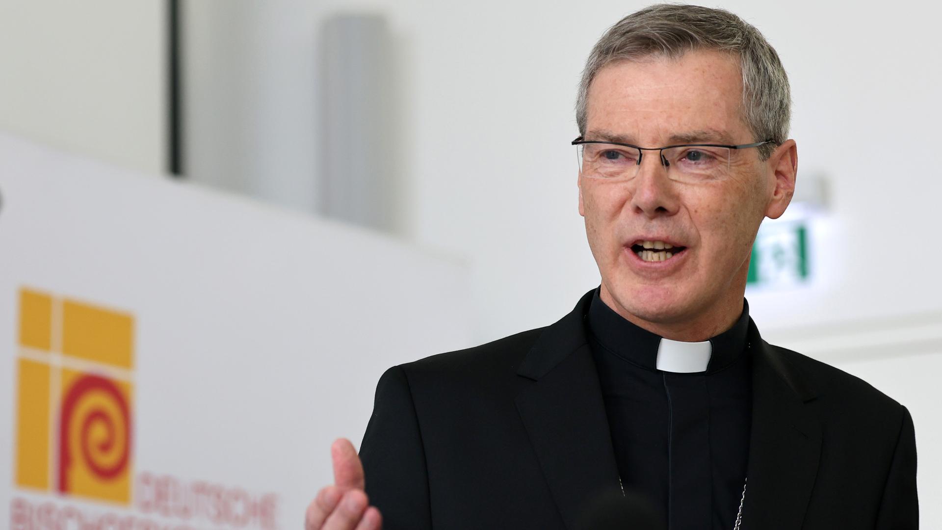 Bischof Heiner Wilmer steht in schwarzem Talar vor dem Logo der Deutschen Bischofskonferenz. Bischof Heiner Wilmer steht in schwarzem Talar vor dem Logo der Deutschen Bischofskonferenz.
