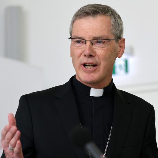 Bischof Heiner Wilmer steht in schwarzem Talar vor dem Logo der Deutschen Bischofskonferenz.