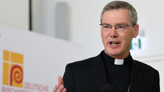 Bischof Heiner Wilmer steht in schwarzem Talar vor dem Logo der Deutschen Bischofskonferenz.