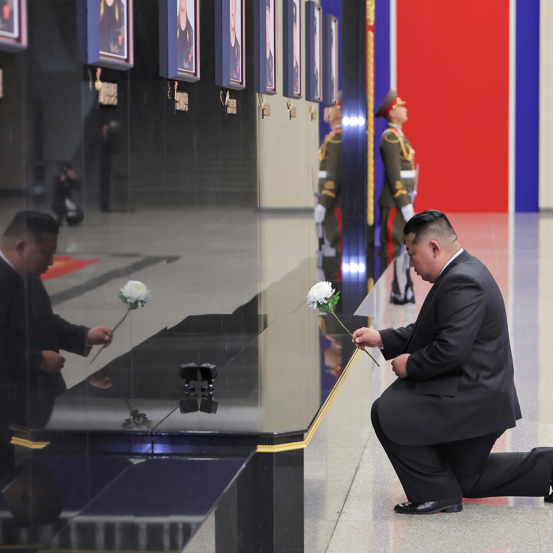 Kim Jong Un kniet vor einer Wand mit Fotos von getöteten Soldaten. In der Hand hält er eine Blume. 