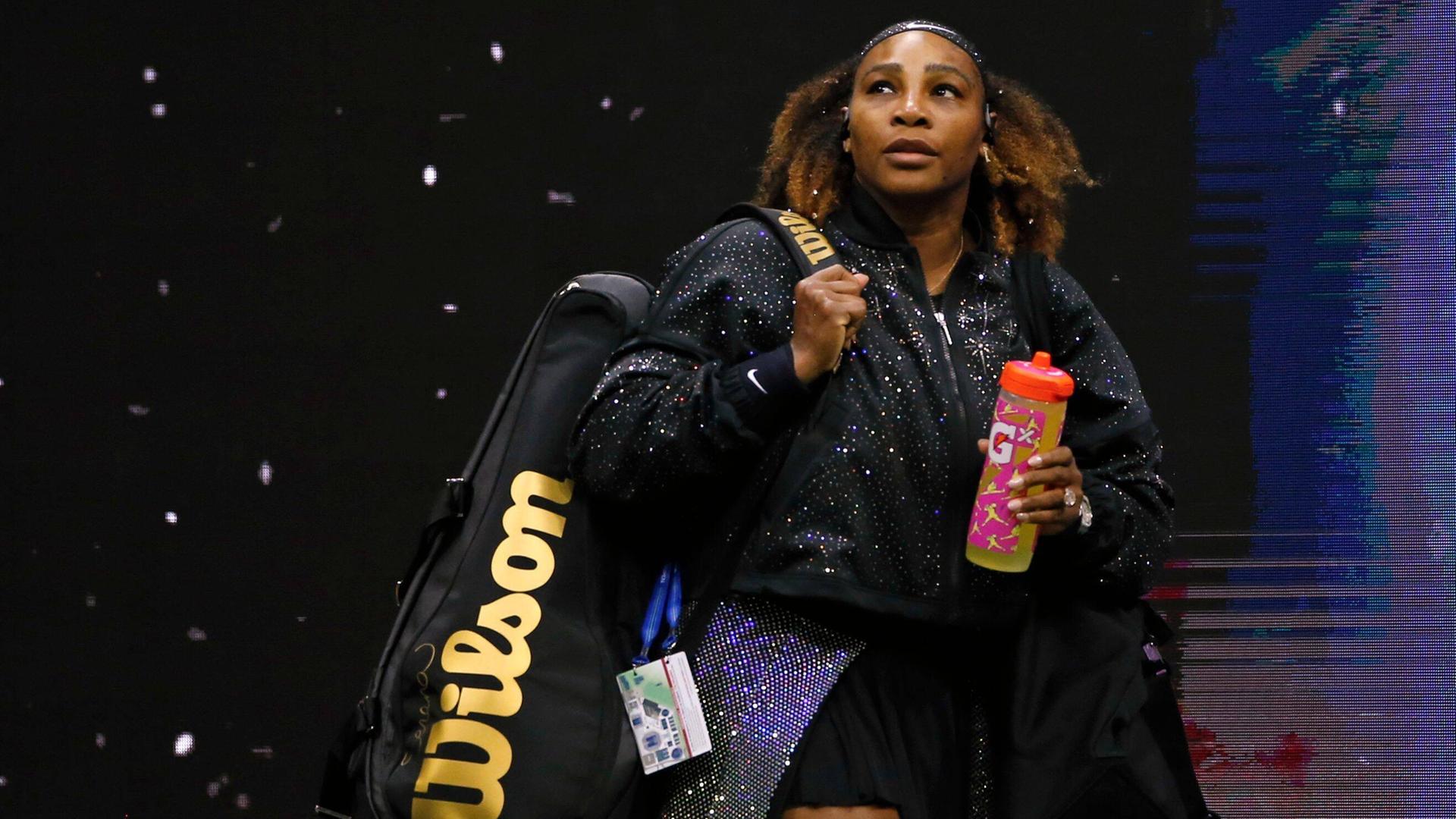 Serena Williams betritt den Tennisplatz in einem schwarzen Dress mit Glitzerumhang, eine große Sporttasche geschultert, mit einer bunten Trinkflasche in der linken Hand.