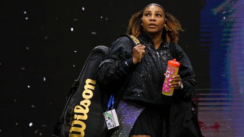 Serena Williams betritt den Tennisplatz in einem schwarzen Dress mit Glitzerumhang, eine große Sporttasche geschultert, mit einer bunten Trinkflasche in der linken Hand. Serena Williams betritt den Tennisplatz in einem schwarzen Dress mit Glitzerumhang, eine große Sporttasche geschultert, mit einer bunten Trinkflasche in der linken Hand.