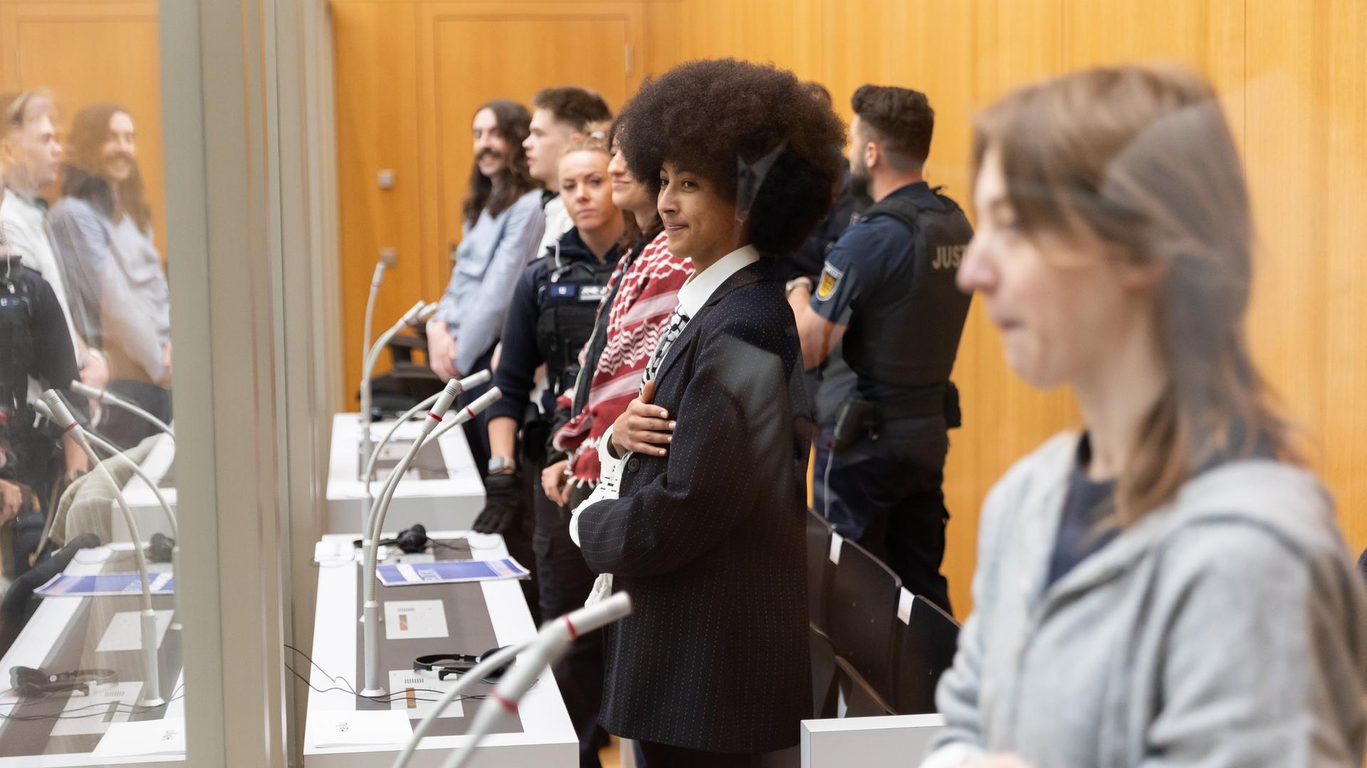 Fünf Menschen stehen als Angeklagte im Gerichtssaal. Sie stehen nebeneinander. Auch mehrere Polizisten stehen dabei. 