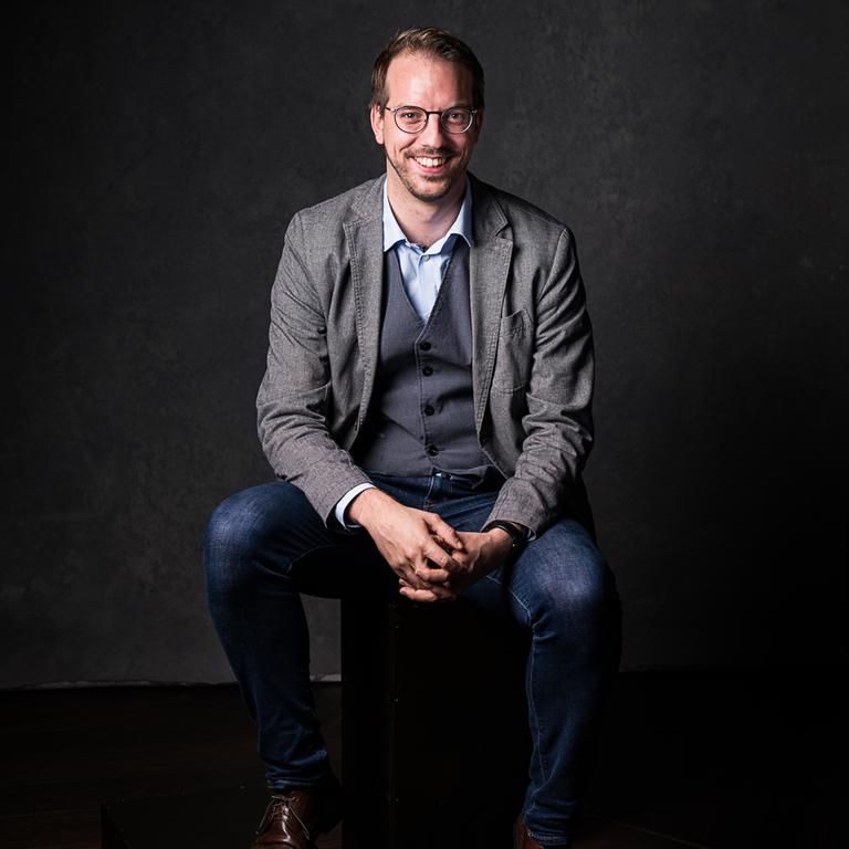 Bibliothekar Marius Müller sitzt im grauen Anzug  auf einem Stuhl vor schwarzen Hintergrund und blickt in die Kamera.