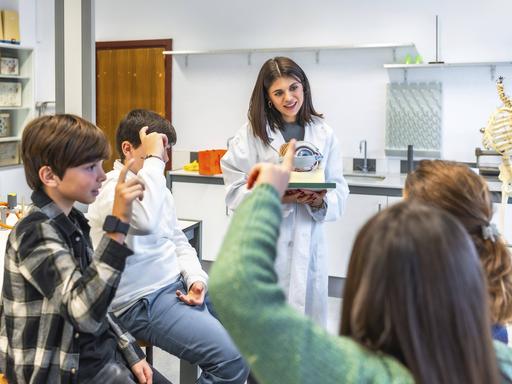 Naturwissenschaftslehrer, der Oberstufenschülern die Anatomie des menschlichen Auges anhand eines Modells während einer Biologiestunde im Schullabor erklärt. Zwei Jungs und zwei Mädchen hören gespannt zu und melden sich.