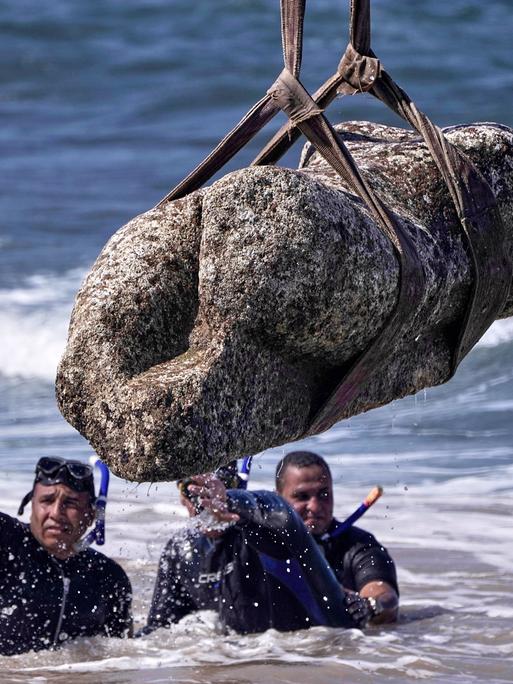 Mehrere Taucher im Wasser. Über ihnen hängt eine Statue an einem Seil.