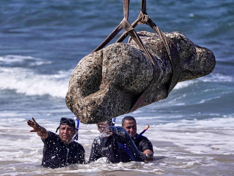 Mehrere Taucher im Wasser. Über ihnen hängt eine Statue an einem Seil.