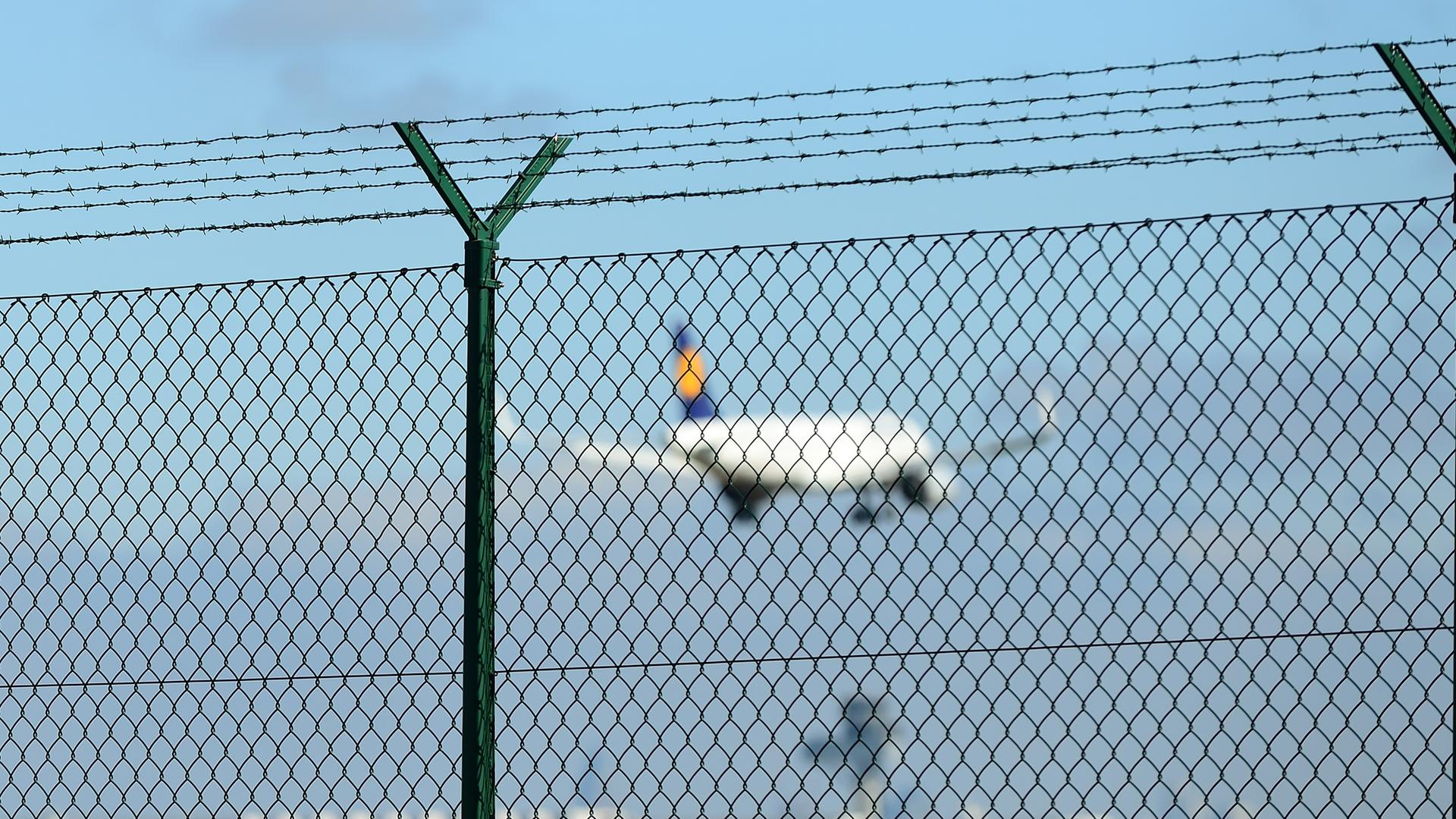 Ein Lufthansa-Flugzeug startet in den Himmel, davor ein Metallzaun. 