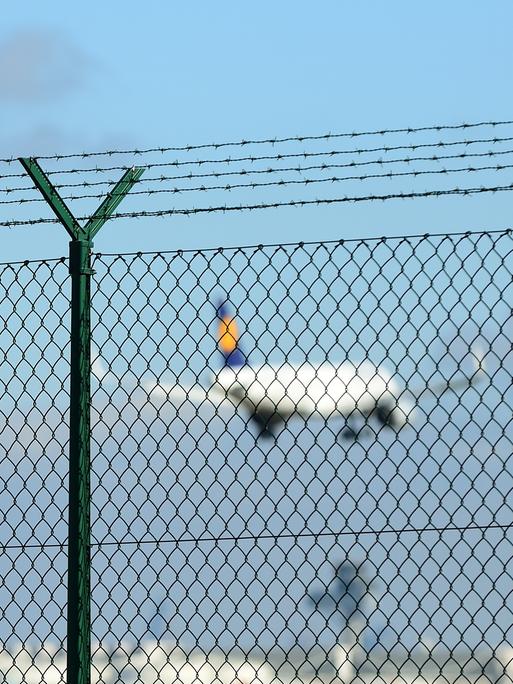 Ein Lufthansa-Flugzeug startet in den Himmel, davor ein Metallzaun. 