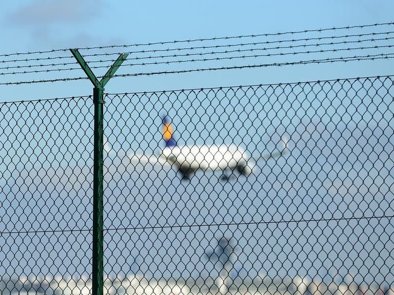 Ein Lufthansa-Flugzeug startet in den Himmel, davor ein Metallzaun. 