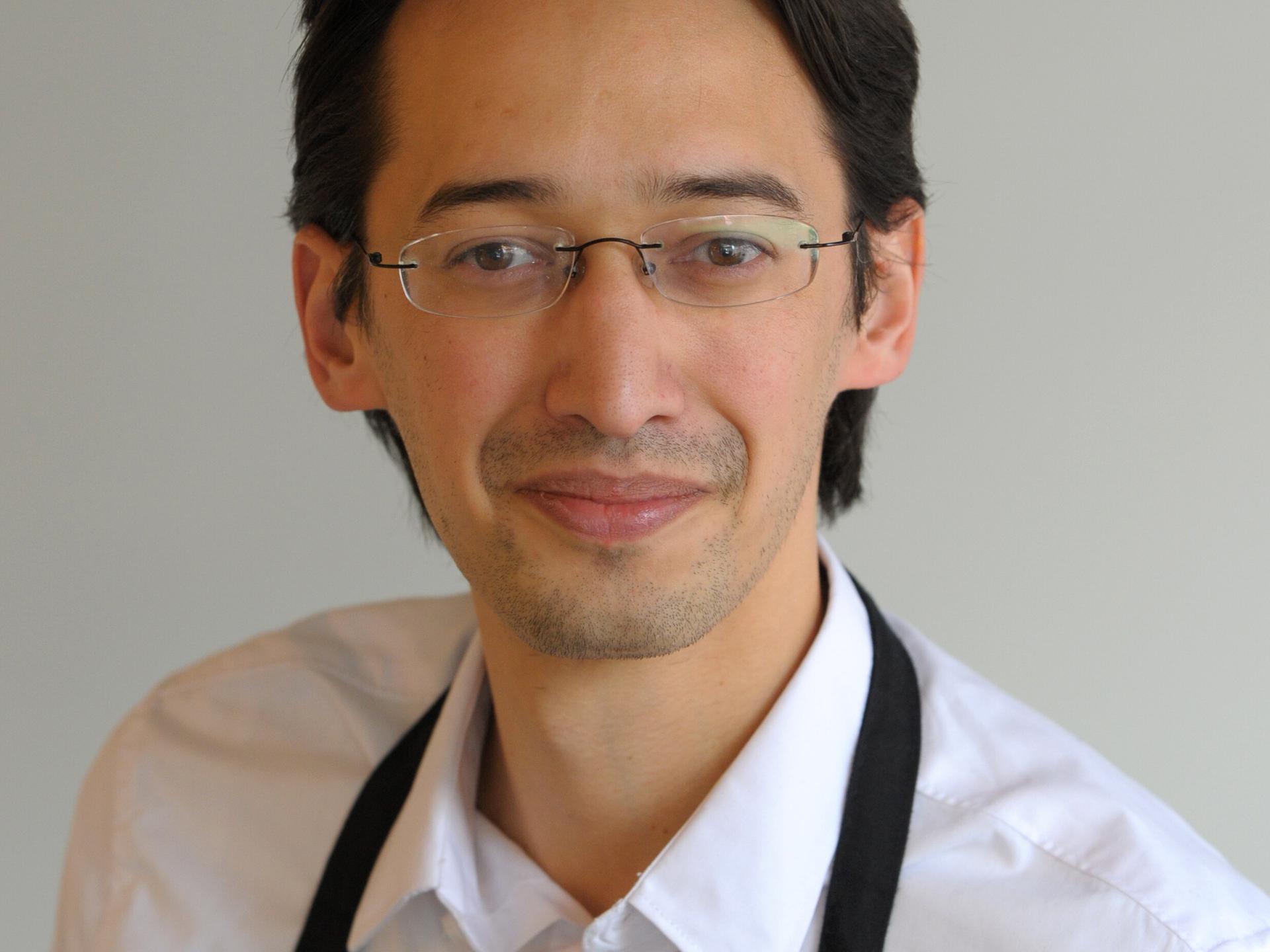 Ramin Goo trägt ein weißes Hemd und schwarze Schürze. Er schaut freundlich in die Kamera bei dieser Porträtaufnahme 