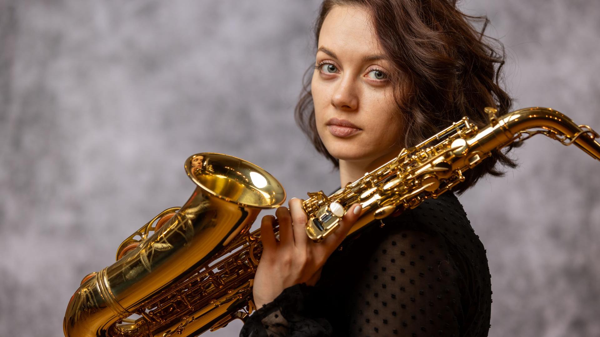 Eine junge Frau mit braunen Haaren dreht ihren Kopf seitwärts und blickt in die Kamera. In ihrer linken Hand hält sie ein Saxofon, das Instrument lehnt auf ihrer linken Schulter.
