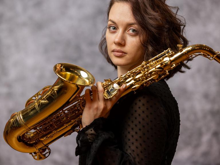 Eine junge Frau mit braunen Haaren dreht ihren Kopf seitwärts und blickt in die Kamera. In ihrer linken Hand hält sie ein Saxofon, das Instrument lehnt auf ihrer linken Schulter.