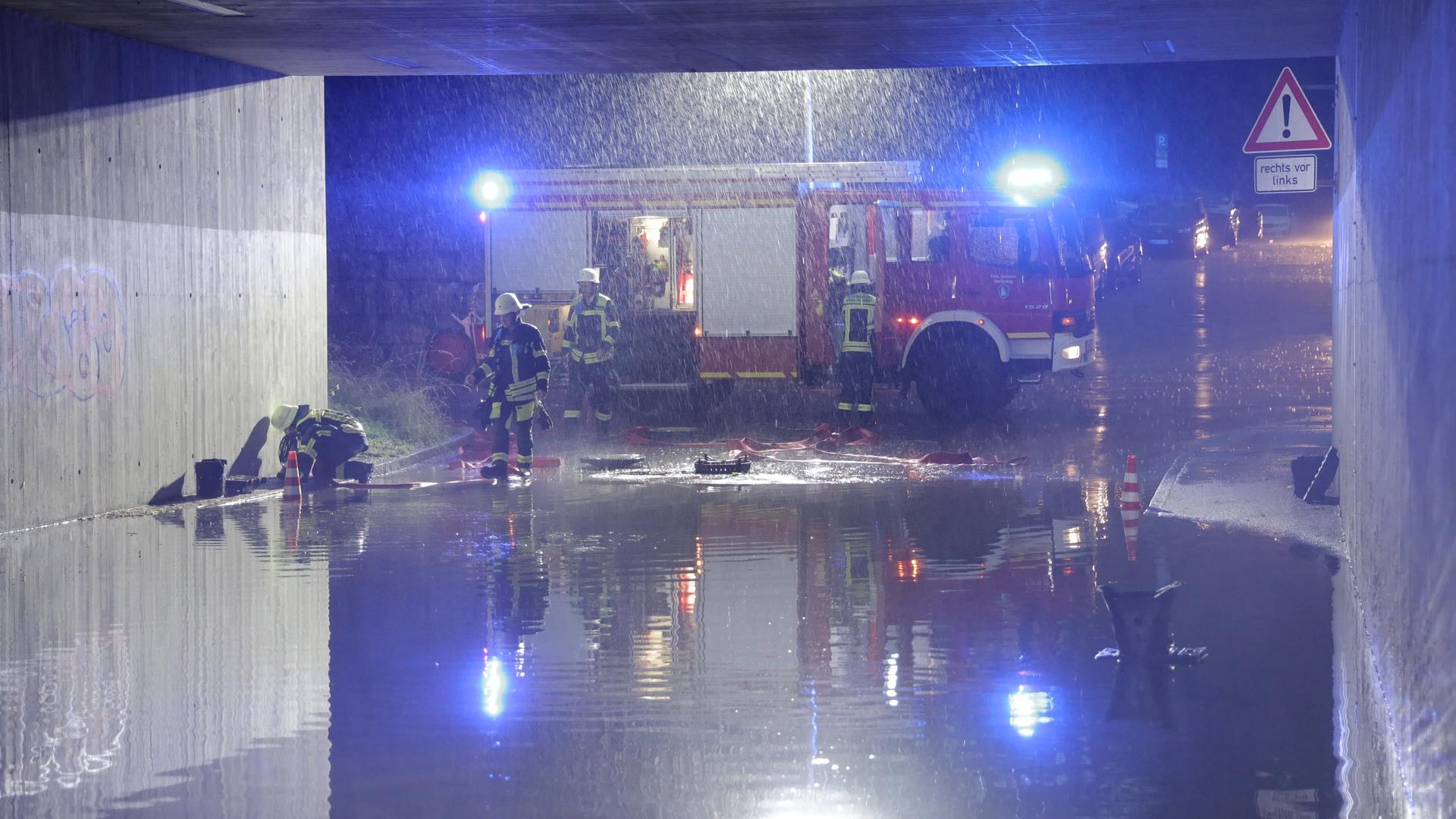 Eine Unterführung steht voller Wasser. Dahinter ein Feuerwehrauto und Feuerwehrleute. Es regnet.