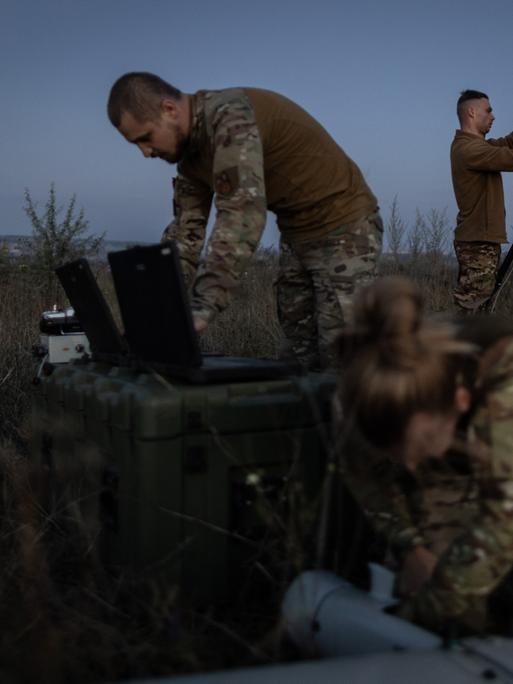 Ein ukrainischer Drohnentrupp bereitet eine Übung mit einer Leleka-Drohne vor. Zwei männliche und eine weibliche Soldatin im Feld bei Dämmerung.