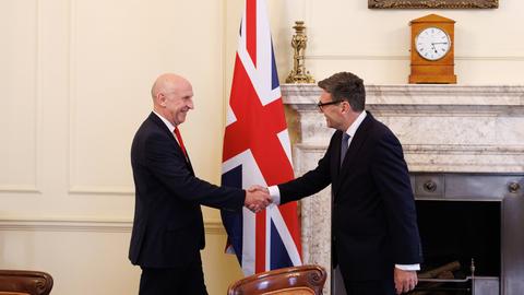 Premierminister Andy Burnham (r.) empfängt in der Downing Street 10 den neuen Finanzminister Healey. Beide stehen vor einem Kamin und reichen sich die Hand.