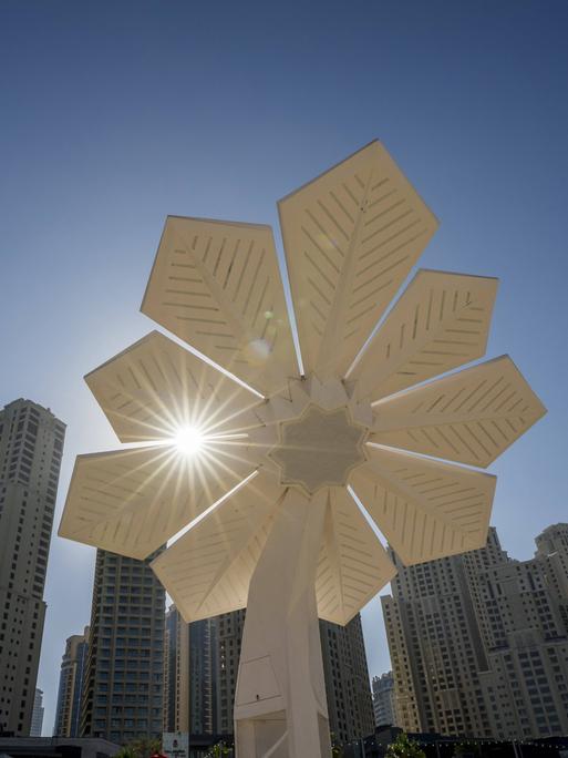 Gegenlicht-Aufnahme: Blumenförmiges Solarpanel vor Wolkenkratzern und wolkenlosem Himmel in Dubai.