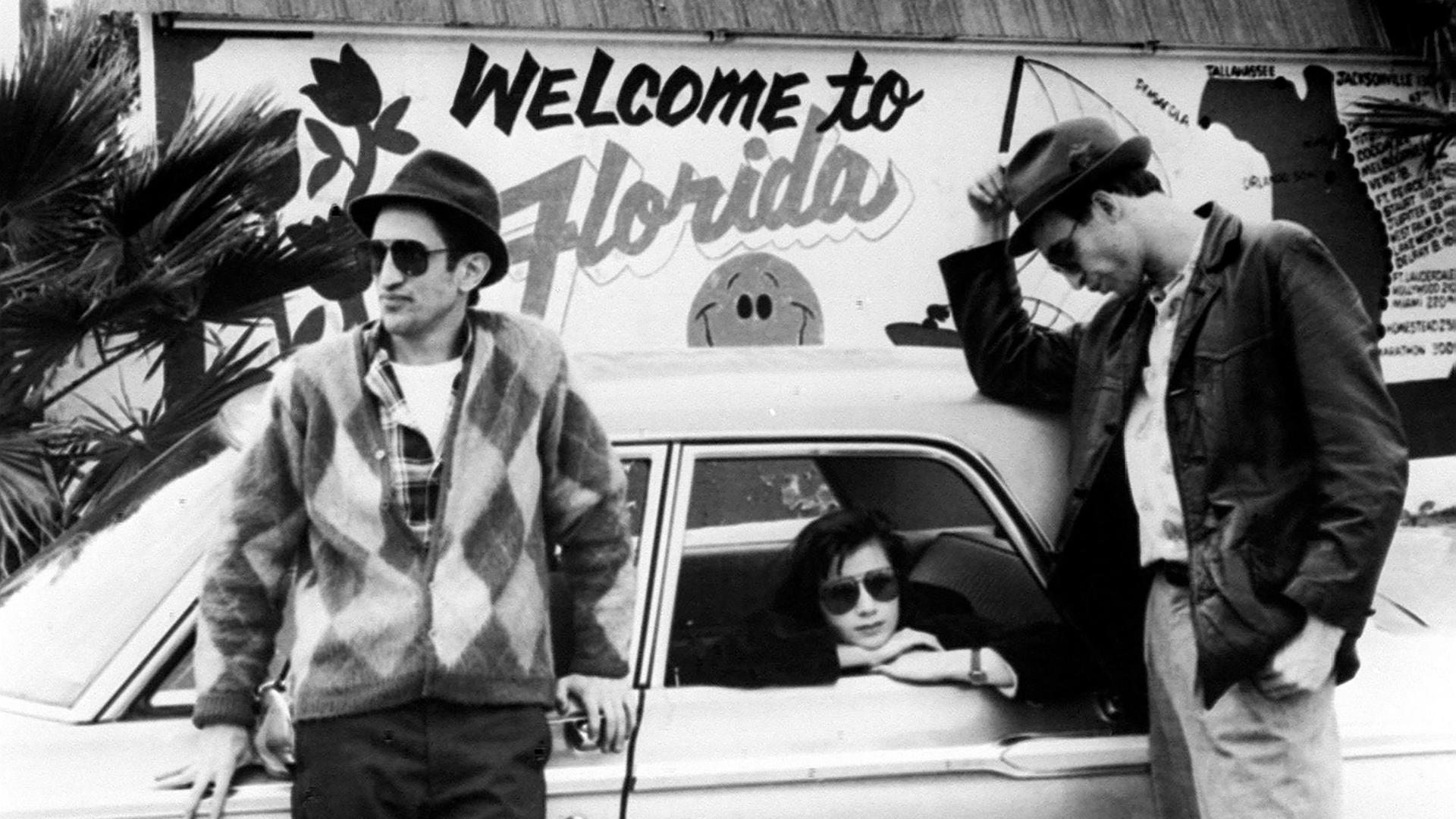 Zwei Männer lehnen an einem Auto, in dem eine Frau sitzt und aus dem Fenster schaut. Im Hintergrund steht auf einer Wand "Welcome to Florida"
