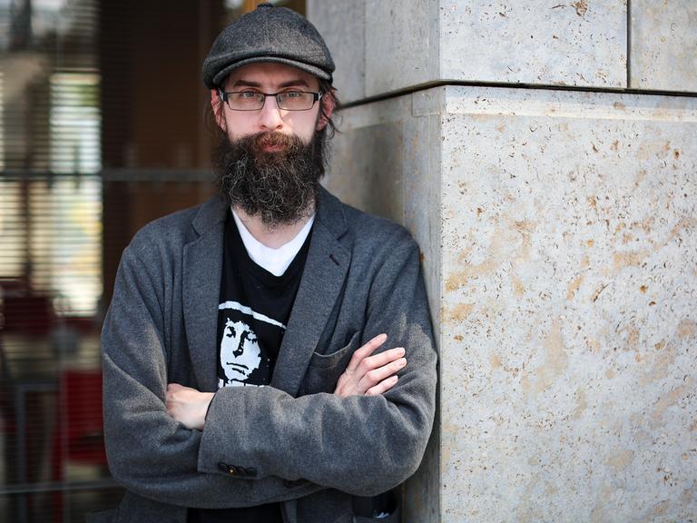 Der Autor Clemens J. Setz steht auf der Leipziger Buchmesse mit verschränkten Armen an eine Mauer gelehnt und blickt in die Kamera.