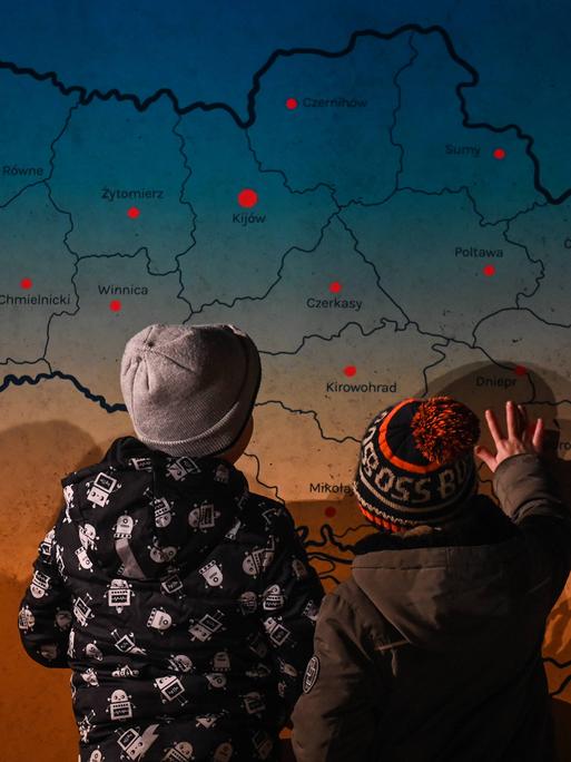 Zwei Kinder sind von hinten vor einer Karte der Ukraine zu sehen, sie zeigen auf die Karte