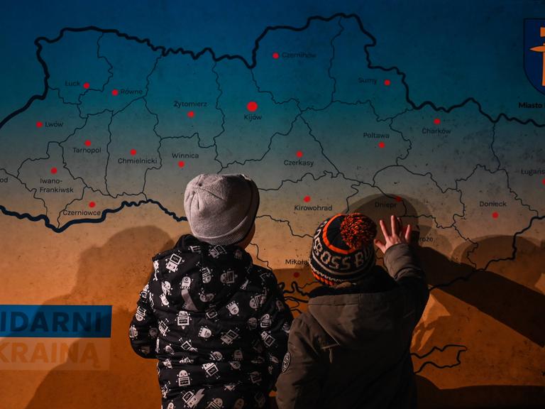 Zwei Kinder sind von hinten vor einer Karte der Ukraine zu sehen, sie zeigen auf die Karte